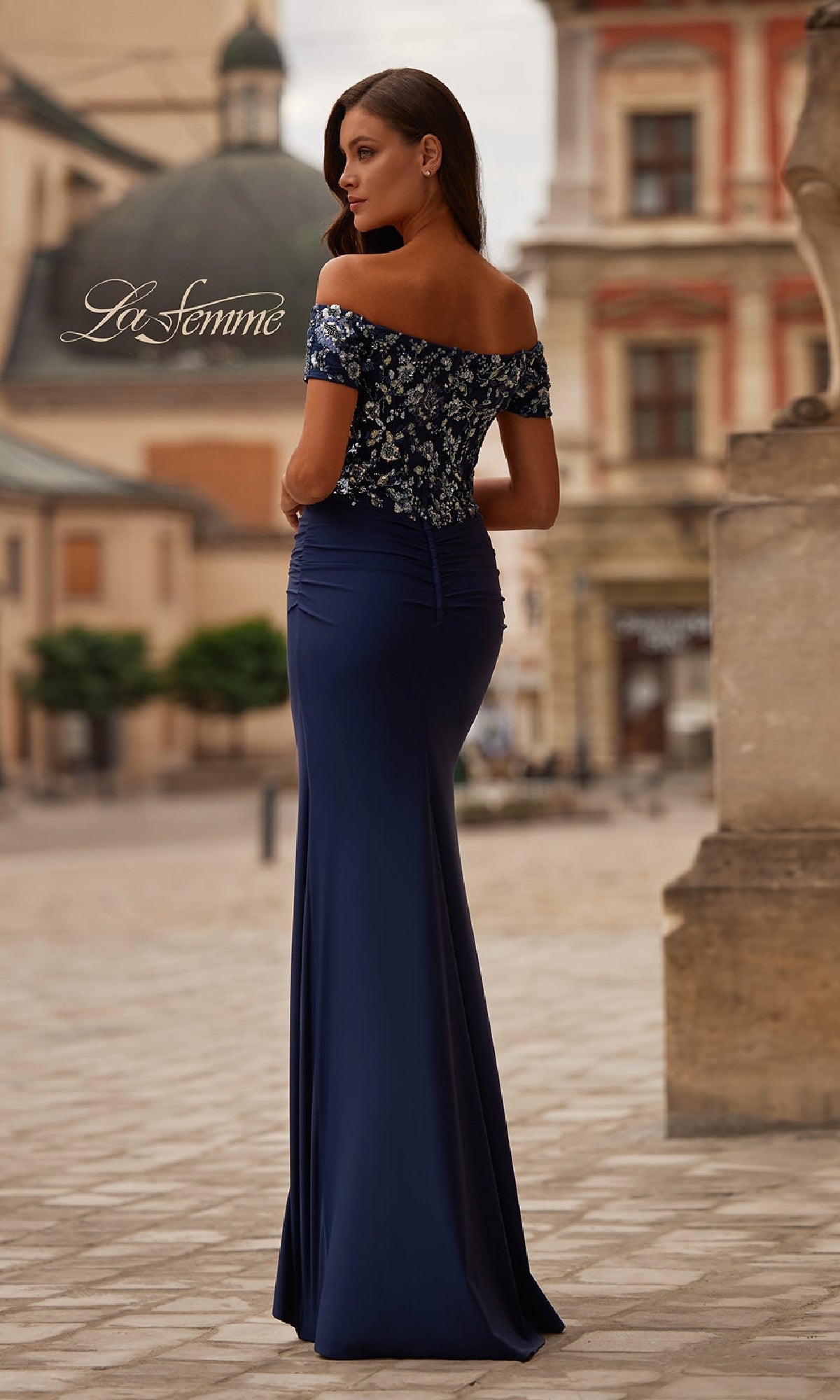 A woman in the Sequin-Bodice Long Navy Prom Dress (La Femme 32739), a fitted off-the-shoulder gown with a sequin bodice, stands outdoors on a stone-paved street, facing away from the camera.